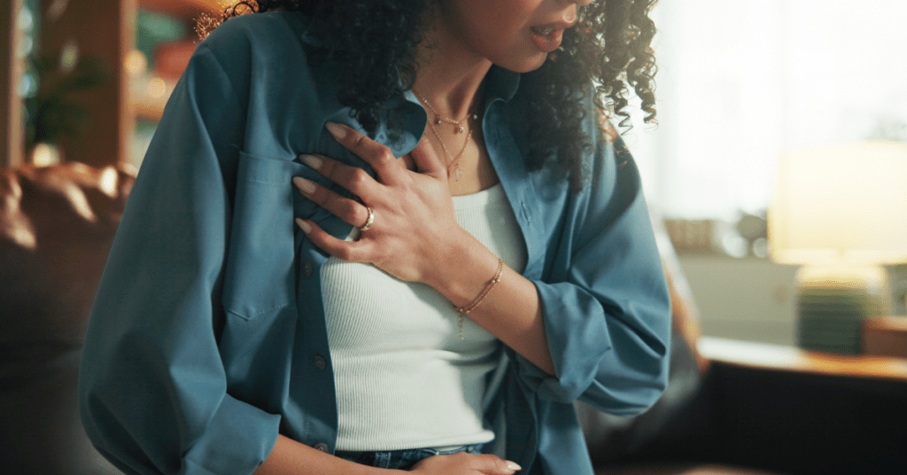 Woman sitting indoors with hand pressed against her chest, appearing to experience stress or chest discomfort.