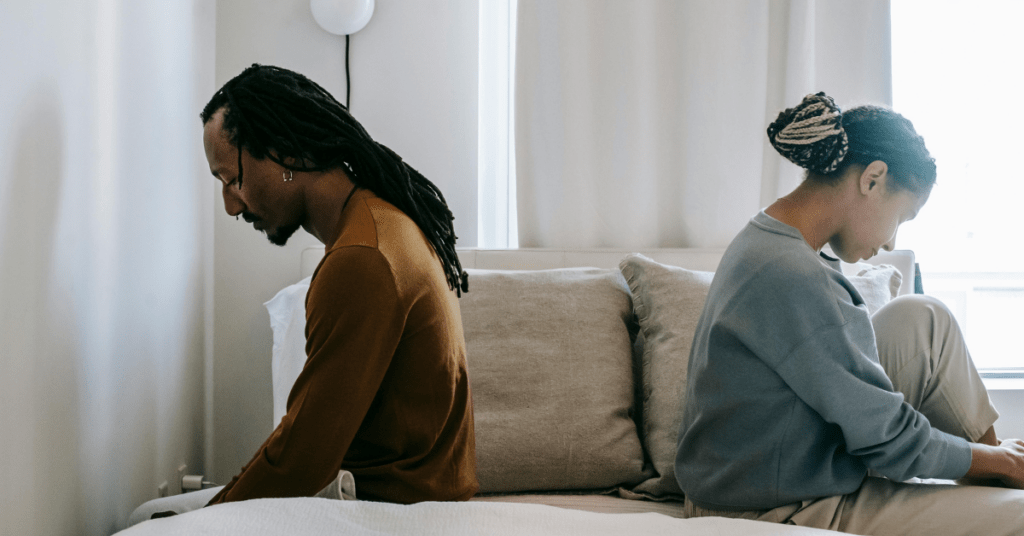 Two people sitting back to back on a couch, looking down and away from each other, suggesting relationship conflict or disconnection