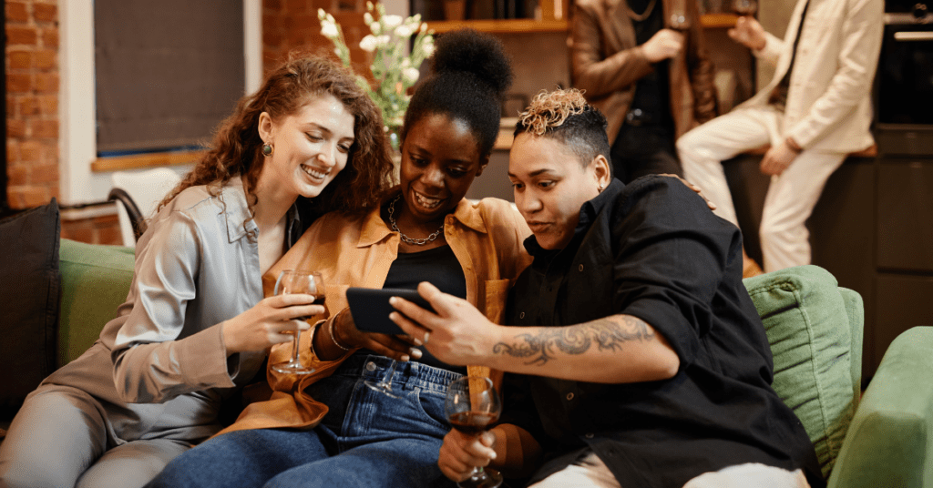 Three queer friends sitting together on a couch, smiling and looking at a phone in a relaxed social setting during an evening gathering