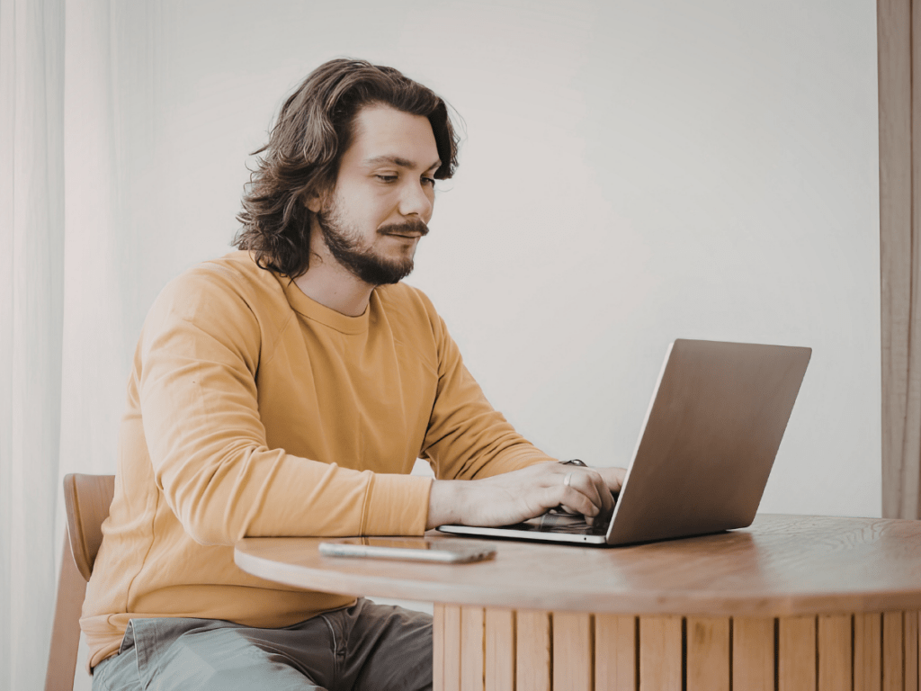 Man in a casual sweater typing on a laptop while sitting at a wooden table at home