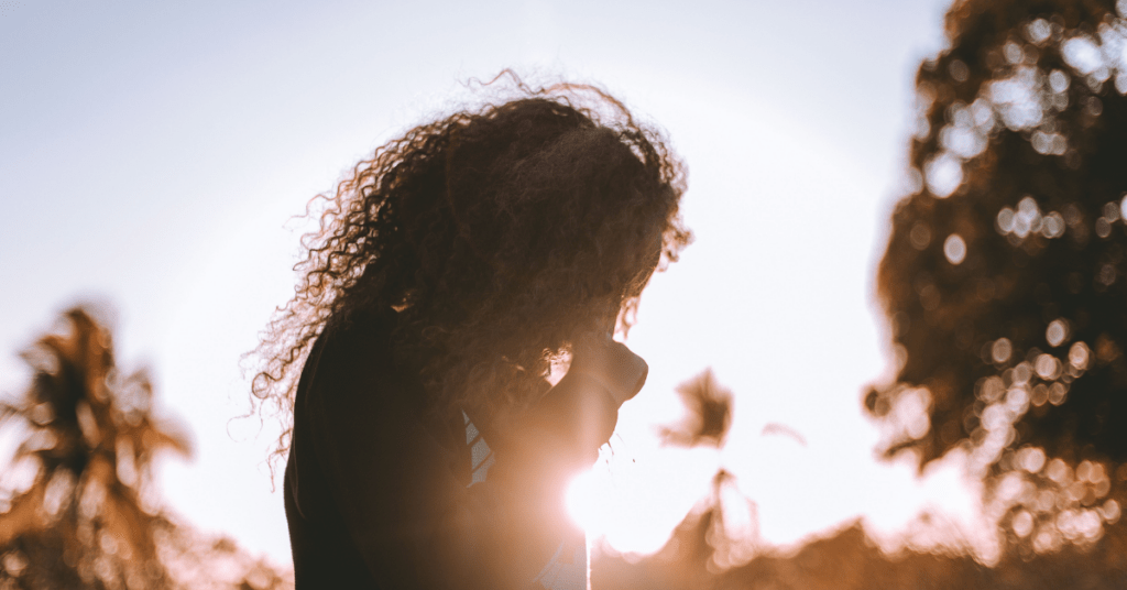 Person with curly hair standing in warm sunlight, reflecting quietly outdoors