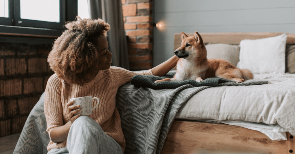 Person sitting beside a dog on a bed while holding a mug in a softly lit bedroom.