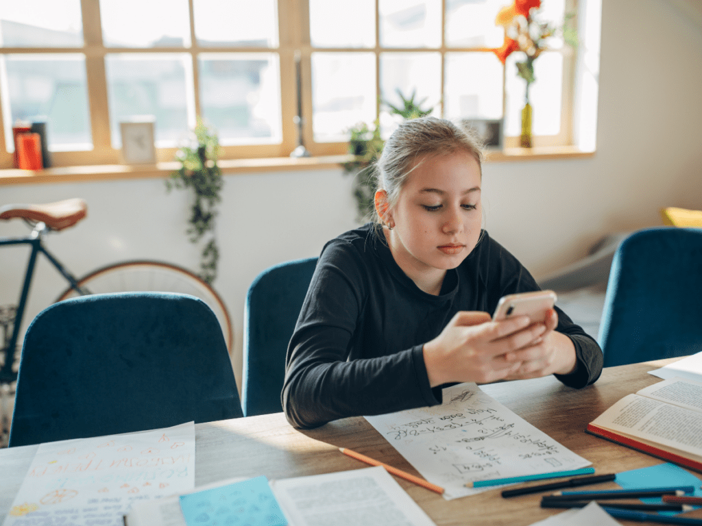Teenager sitting at a table looking at a phone while homework and papers sit nearby