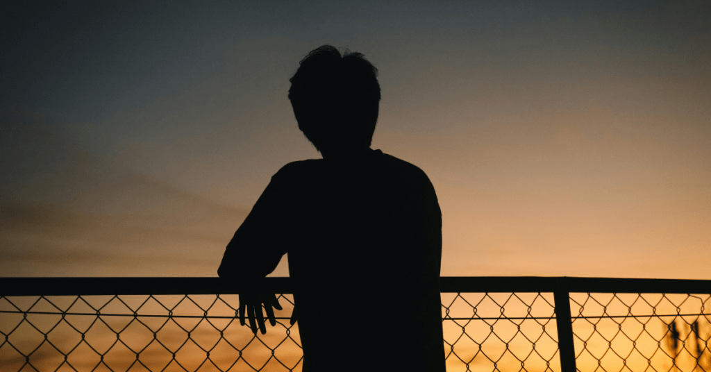 Silhouette of a person standing at a chain-link fence during sunset, looking out toward the horizon.